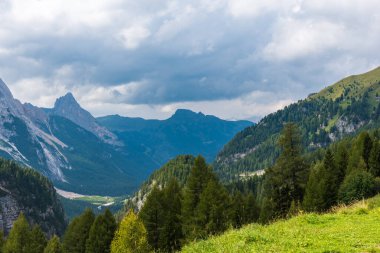 Parlak bulutlu gökyüzünün altındaki yeşil ormanlarla kaplı Dolomitler.