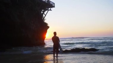 Beyaz bir adam bir koyda sörf tahtası ile okyanus tarafından standları ve mesafe içine güzel bir gün batımı görünüyor. Güneş bir kayanın arkasından bakıyor ve suya yansıyan.