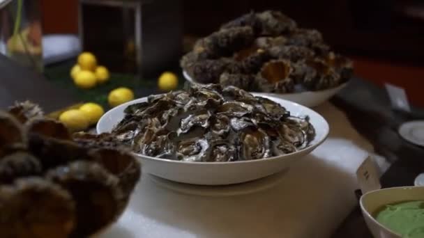 在柜台上的白色盘子上的牡蛎特写镜头 旁边是煮熟的海胆盘 后面是两个花瓶的花和柠檬 全景拍摄 — 图库视频影像