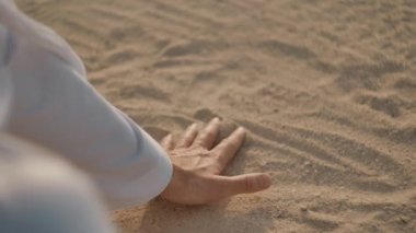 Beyaz giysili beyaz adam elini kumların üzerinden çalışır, bir yumruk içine bir avuç alır, sonra onun el ve kum dökülen onun parmaklarının arasından açar. Güneş parlıyor.