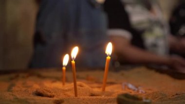 Üç sarı yanan mum kumun üzerinde durmak. Mum alevi. Sert balmumu kumun üzerinde görünür. Arka plan - insanlar. Loş mum ışığı. Tapınağın mumlar.