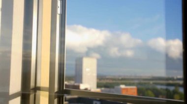 Beyaz gömlekli bir adam pencereye yürüyor ve sağ elini yükseltir, içinde siyah bir telefon var. Güneşli, parlak havalarda yukarıdakinden şehir manzarasına bakan pencereden. Zemin pencereleri