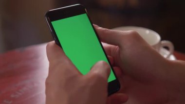Masada oturan ve telefon tutan bir adamın elini yakın çekim. Bir adam parmaklarını ekrana karşı yönetiyor. Ekranda yeşil bir arka plan vardır. Bir kafe atmosferi etrafında.