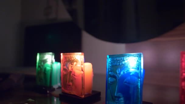 桌子上有五颜六色的蜡烛 前面是长方形的玻璃面具 上面有一个女人的脸 眼睛紧闭 反映了蜡烛的颜色和火焰的光芒 — 图库视频影像