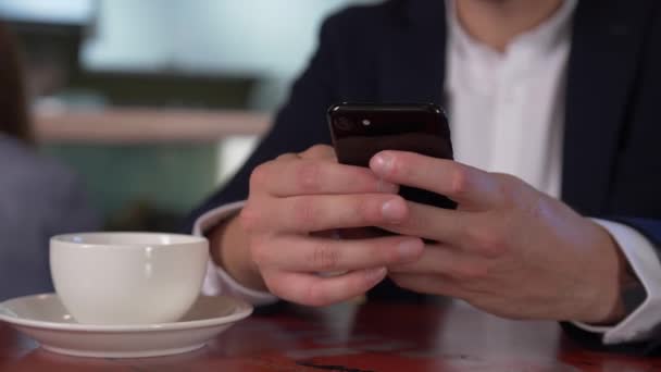 一名身穿白衬衫和蓝色夹克的男子坐在一张小桌旁 在手机上表演 桌子上有一杯茶 他拿着它喝了一口 可以看出 一个人有胡子 — 图库视频影像