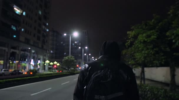 那个穿着黑色皮夹克和科皮松的家伙背着一个背包 正沿着夜城的街道走着 街上的灯光闪闪发光 — 图库视频影像