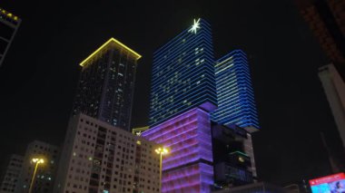 Macau gece hayatı. Sarı, pembe ve mavi ile parıldayan güzel yüksek binaların aşağıdan yukarıya görünümü. Bazı pencereler yanıyor. Fenerler sokağa iner..