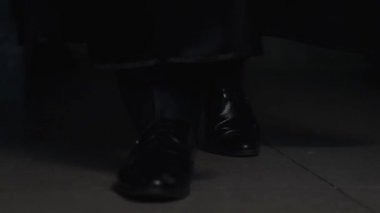 Siyah pelerinli, pantolonlu ve deri çizmeli bir adam gri odadaki fayansların üzerinde yavaşça yürüyor. Oda alacakaranlık. Bacakları yakın çekim. Ön görünüm.
