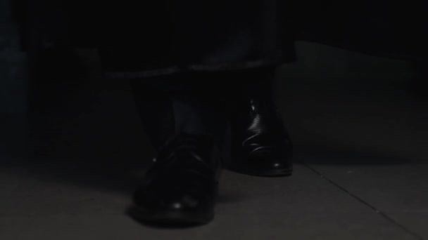 一个穿着黑色斗篷 裤子和皮靴 脚上有鞋带的男人慢慢地走在灰色房间的瓷砖上 房间里一片暮色 腿的特写 — 图库视频影像