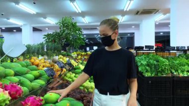 Siyah tişörtlü, beyaz pantolonlu ve yüzünde tıbbi maske olan sarışın, beyaz bir kız marketin tezgahından meyve seçiyor. Olgun, raflarda farklı meyveler. Covid-19 sırasında alışveriş..