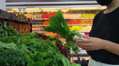 Siyah tişörtlü, beyaz pantolonlu ve yüzünde sağlık maskesi olan sarışın beyaz bir kız marketin tezgahından yeşillikleri seçiyor ve bir sürü yeşil salataya bakıyor. Covida-19 'da alışveriş..