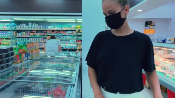 一个带着医疗面具的金发白人女孩走到冰箱前 打开冰箱 拿起一块肉 看了看 毫无选择地把它拿了回来 Covida 19期间的购物 — 图库视频影像