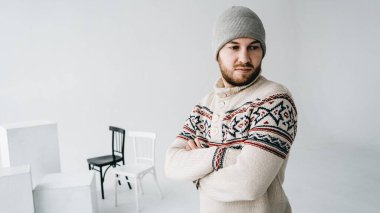 Bıyıklı, keçi sakallı, gri şapkalı, beyaz örgülü, dar kazaklı beyaz bir adam kollarını geometrik şekiller ve sandalyelerle ışık odasında kavuşturuyor. Yan bakış