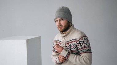 Keçi sakallı, bıyıklı, gri şapkalı ve desenli beyaz örülü süveter giyen beyaz bir beyaz adam. Parlak bir odada elinde kazak koluyla duran bir adam var.