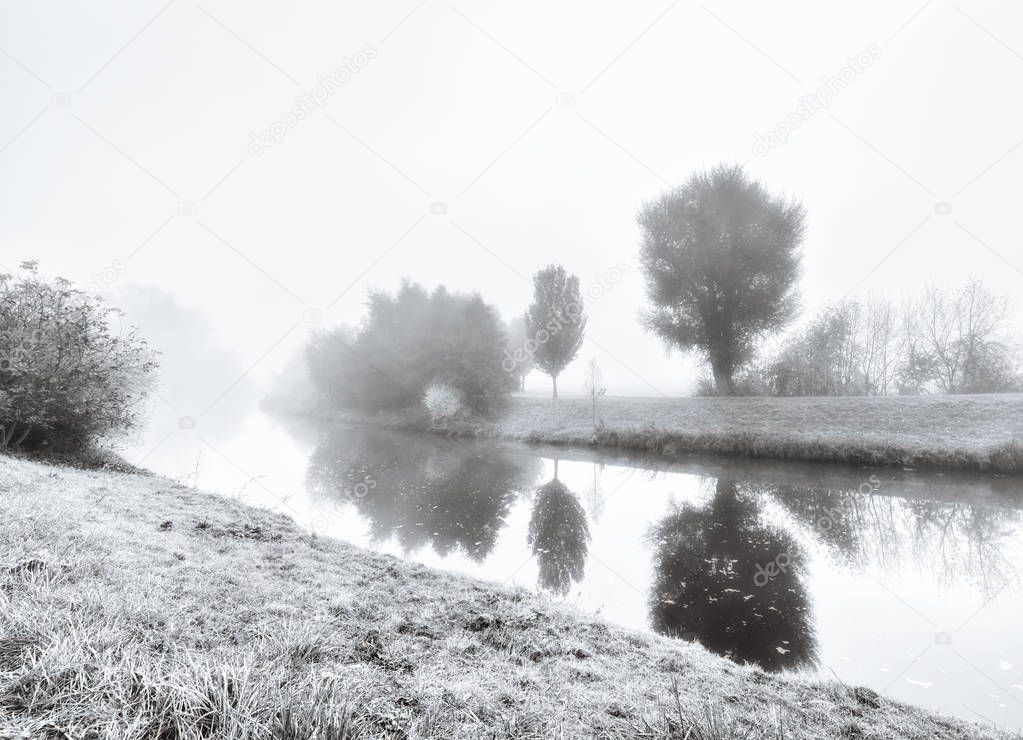 Black and white monochrome outdoor image taken at a river  with hoar frost and trees reflecting on the water on foggy autumn or winter morning, a promenade and a bench to rest