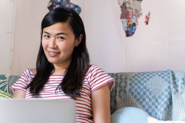 Asyalı uzun saçlı bir kadın Camara 'ya bakıyor, oturma odasında laptopuyla çalışıyor. portre,