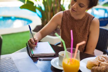 Bahçede terasta oturan bir kadın, elinde bir havuz, masada bir bilgisayar ve kahvaltı ile bir not defterine yazıyor.