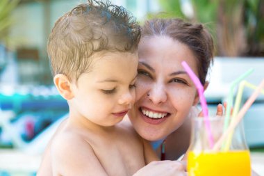 Islak saçlı ve havuzda bir bardak meyve suyu içen bir çocuğun yanında mutlu bir ifadeyle bir kadının yüzü.