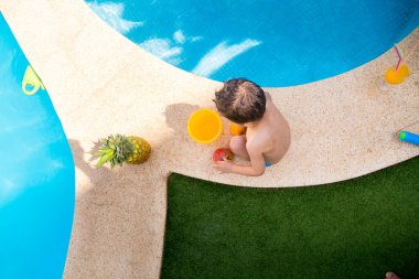 Havuzda farklı bir meyve çeşidi ve havuzun kenarında çimlerle çevrili bir bardak portakal suyu olan beyaz bir çocuğun havadan çekilmiş fotoğrafı.