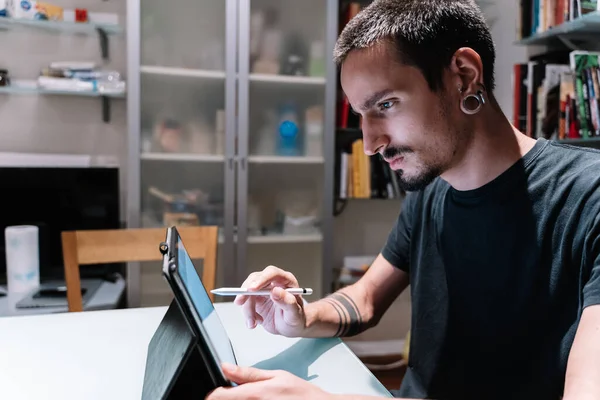 Profile of a man with piercings, beard and moustache using a tablet with a digital pen in a room full of books