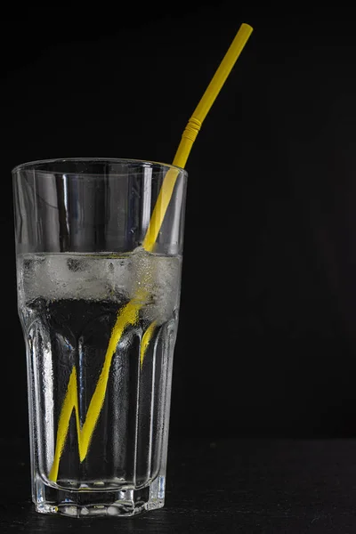 Glass cup with water, ice cubes and yellow straw on black background in vertical with copy space