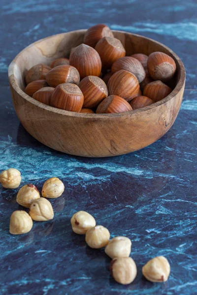 Close-up of peeled hazelnuts and whole hazelnuts in wooden bowl on blue marble background in vertical