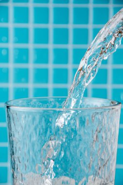 Su jetinin cam bardağa düşüşünün detaylı görüntüsü, açık mavi mozaik arka planda, dikey