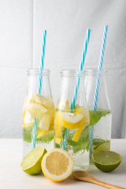Mavi pipetli üç şişe, su ve limon, kireç ve nane dilimleri, beyaz masa üzerinde yarım limon ve tahta kaşık, beyaz arka plan, dikey