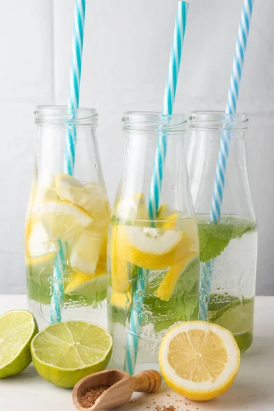 青いわら 水とレモン ライムとミントのスライスを持つ3本のボトルのクローズアップ 半分レモンと茶色の砂糖 白の背景 垂直と木製のスプーンを持つ白いテーブルの上 — ストック写真