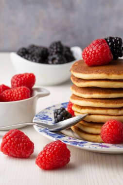 Üzerinde böğürtlen, ahududu ve kaşık bulunan gözleme tabağı beyaz ahşap masa, ahududu ve böğürtlen dolu kaseler, gri arka plan, dikey