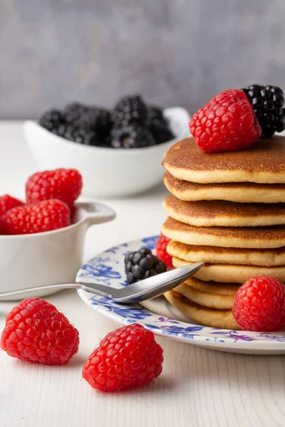 Üzerinde böğürtlen, ahududu ve kaşık bulunan gözleme tabağı beyaz ahşap masa, ahududu ve böğürtlen dolu kaseler, gri arka plan, dikey