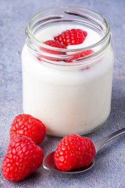 Yakın plan kaşık, ahududu ve cam kavanoz beyaz yoğurt ve gri arka planda ahududu, dikey