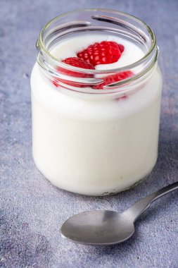 Beyaz yoğurtlu kaşık ve cam kavanoz manzarası ve gri arka planda ahududu, dikey.