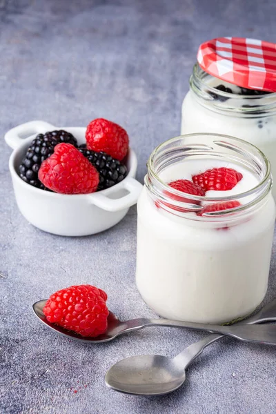 İki kaşığın üst görüntüsü, ahududu, böğürtlen ve ahududu dolu iki kavanoz yoğurt, gri arka planda, dikey, fotokopi alanlı.