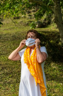 Beyaz ve sarı giyinmiş olgun bir kadın koruyucu bir maske takıyor, bir çayırda dikey, odaklanmamış bir arka planı var.
