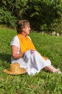 Beyaz ve sarı giyinmiş, elinde koruyucu bir maskeyle çayırda oturan, odaklanmamış, dikey bir geçmişi olan olgun bir kadının yakın çekimi.