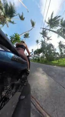 Dikey video. Motosikletli bir kadın, tropikal yoldaki eğri büğrü gölgeli palmiye dalının yanından geçerek, alçak açılı Ada Keşif Konsepti 'ne yakalandı.