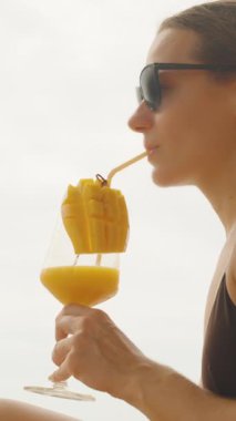 Dikey video. Mayolu, saç örgülü ve güneş gözlüklü bir kadın pipetle sallanırken kumsalda, yan görüşte, gevşemiş tropikal yaşam tarzı konsepti