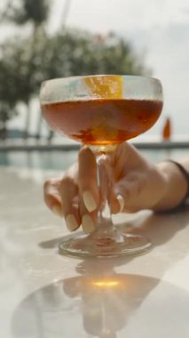 Dikey video. Uzun tırnaklı bir kadın havuz kenarında kırmızı sıvıyla dolu bir kokteyl bardağı tutuyor. Parlak gün ışığında. Yakın çekim.