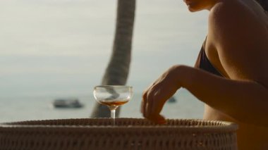 Bikinili ve güneş gözlüklü kadın, gün batımında okyanusa bakarken kokteyl içerler. Yumuşak ışık ve palmiye gövdesiyle yan yana dururlar. Tatil Gevşemesi.
