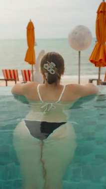 Dikey video. Bikini 'deki Kadın Gözlerinin Arkasından Yakaladığı Orange Şemsiyeleriyle Okyanusa Karşı Sonsuz Havuz Kenarı' nda Dinlenen Kollar