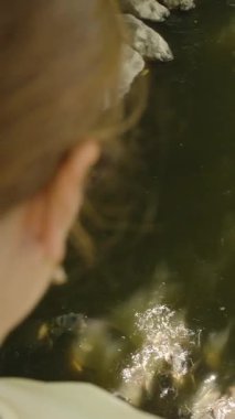 Dikey video. Arkadan atılan peletleri nazikçe dalgalanma ve gölgeli yansımalarla göle atan kadın, doğayla etkileşimde.