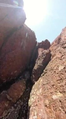 Dikey video. Parlak güneş ışığı altında kayan alt kamerada küçük su akıntısı olan kırmızımsı kahverengi kayalar arasındaki dar aralığı kapat