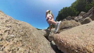 Güneşli bir günde kıyı şeridi yakınlarındaki kayalara tırmanan kadın hareketini ve kayalık patika macerasını ve açık hava keşiflerini düşük açıyla gösteriyor.