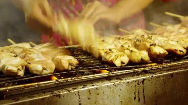 Açık ızgarada pişen tavuk şişleri, yükselen dumanla ve satıcı eliyle sokak yemekleri tezgahında şiş çeviren ızgara yiyecek kavramını kapatın.
