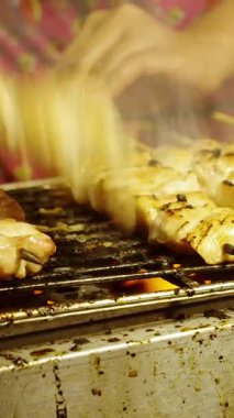 Dikey video. Açık ızgarada pişen tavuk şişleri, yükselen dumanla ve satıcı eliyle sokak yemekleri tezgahında şiş çeviren ızgara yiyecek kavramını kapatın.