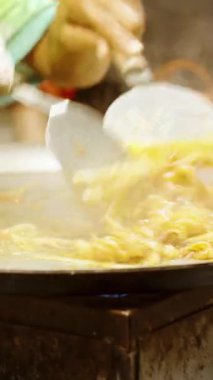 Dikey video. Sokak satıcılarının elleri büyük sıcak tavada makarna ile şehriye karıştırırken Tayland 'daki kalabalık markette buharlı Pad Thai pişiriyorlar.
