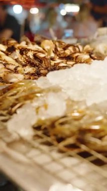 Dikey video. Buz üzerinde şiş karidesleri ve gece pazarında metal askılarda sergilenen ızgara deniz ürünleri, taze deniz ürünleri konsepti.