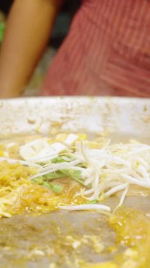 Dikey video. Sokak satıcısını hemen kapatın. Tayland pazarında Pad Thai pişirirken tofu ve fasulye filizlerini büyük sıcak tavada spatulayla karıştırın. Sokak Gıda Konsepti.