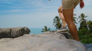 Kayalık bir bakış açısıyla yürüyen, arka planında parlak gün ışığında palmiye ağaçları ve okyanus olan, alçak açılı, çakışan taş Koh Samui konsepti olan kadın.